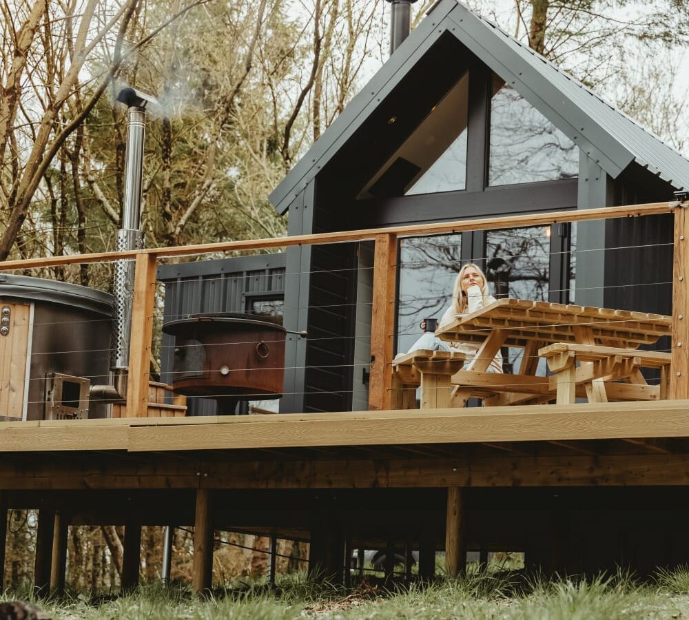 Nestled among the trees, a small cabin with a glass front sits on a wooden deck. This perfect glamping spot features a picnic table and a person enjoying the serene surroundings, embodying the tranquil essence of Porth Eryri.