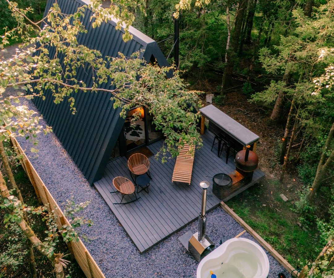 Aerial view of a charming A-frame cabin nestled in a lush forest, perfect for glamping near Porth Eryri. The cabin features a wooden deck with two chairs, a bench, and a freestanding bathtub. The setting is tranquil and surrounded by greenery.