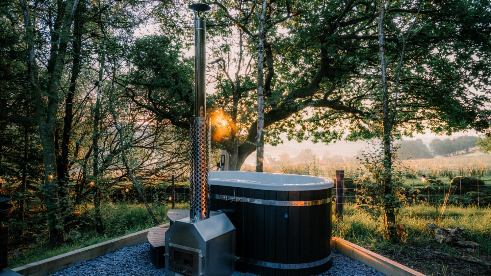 A serene outdoor scene at Porth Eryri Glamping features a hot tub with a tall chimney, set on gravel, surrounded by lush greenery and trees. The sun is setting in the background, casting a warm glow through the branches and creating a peaceful atmosphere.