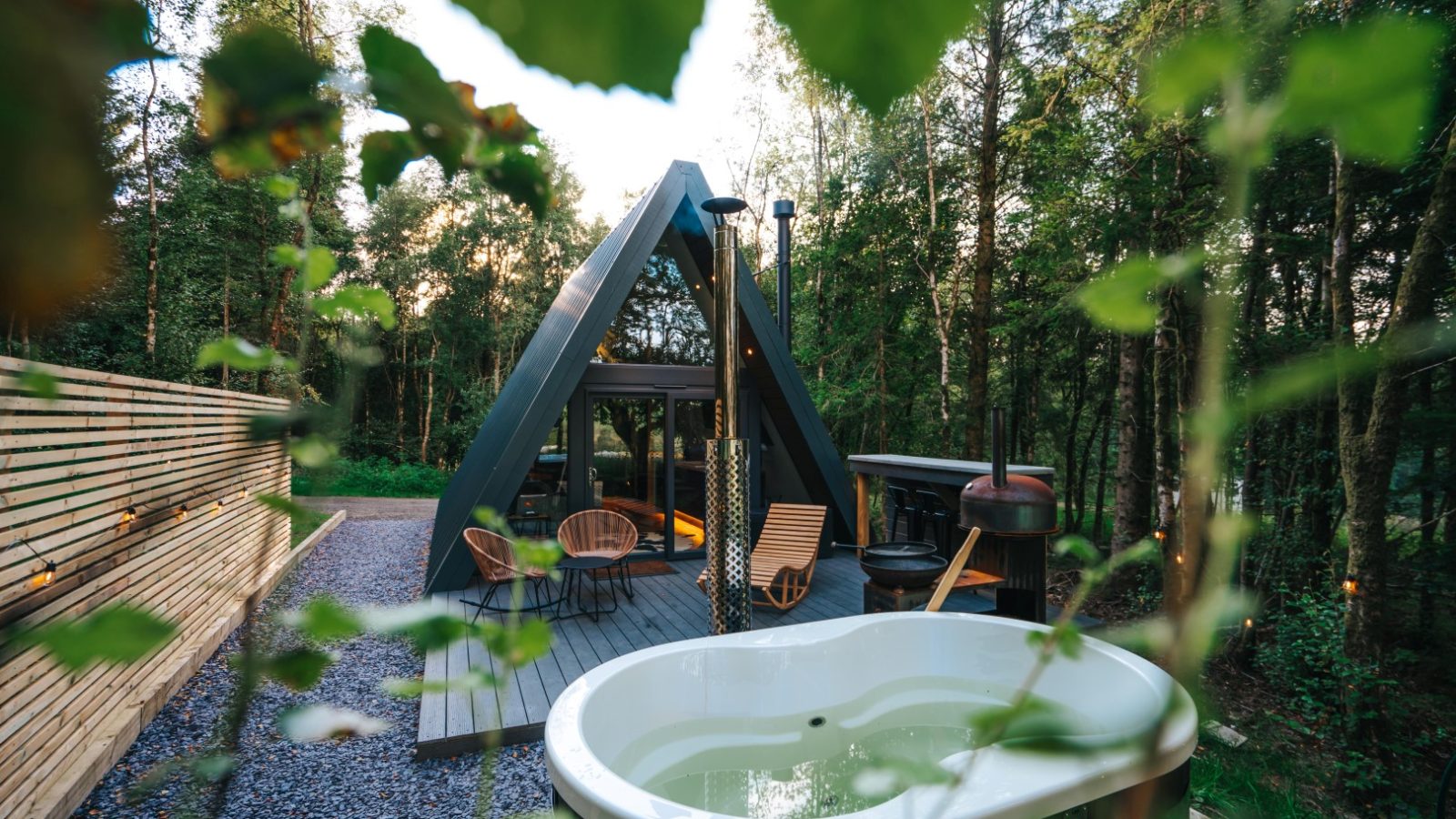 Nestled in Porth Eryri, this cozy A-frame cabin offers a glamping retreat with a hot tub amidst the trees. Wooden deck chairs and a small chimney enhance the deck's charm, while surrounding leaves frame the serene, natural atmosphere.