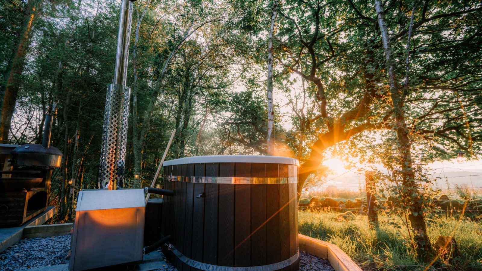 Nestled among the trees of Porth Eryri, a rustic hot tub offers a serene glamping experience. With sunlight streaming through the leaves, its tall chimney and surrounding gravel and wooden planks create a tranquil outdoor retreat.