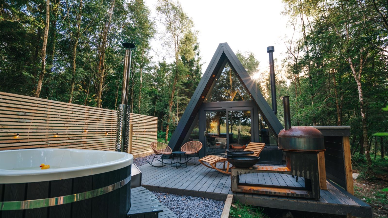 A cozy A-frame cabin nestled in the forest offers a glamping experience with its wooden deck featuring a hot tub, two lounge chairs, a round table, and a small barbecue. Located in Porth Eryri, tall trees surround the area as sunlight dances through the foliage.