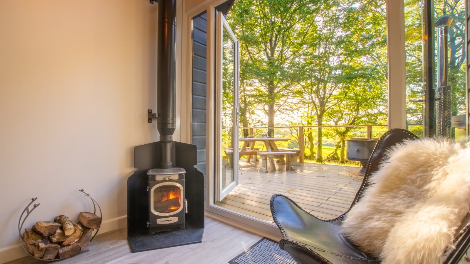 Cozy glamping room with a wood stove, a butterfly chair draped in a fur throw, and views of Porth Eryri's trees with a picnic table on the deck outside.