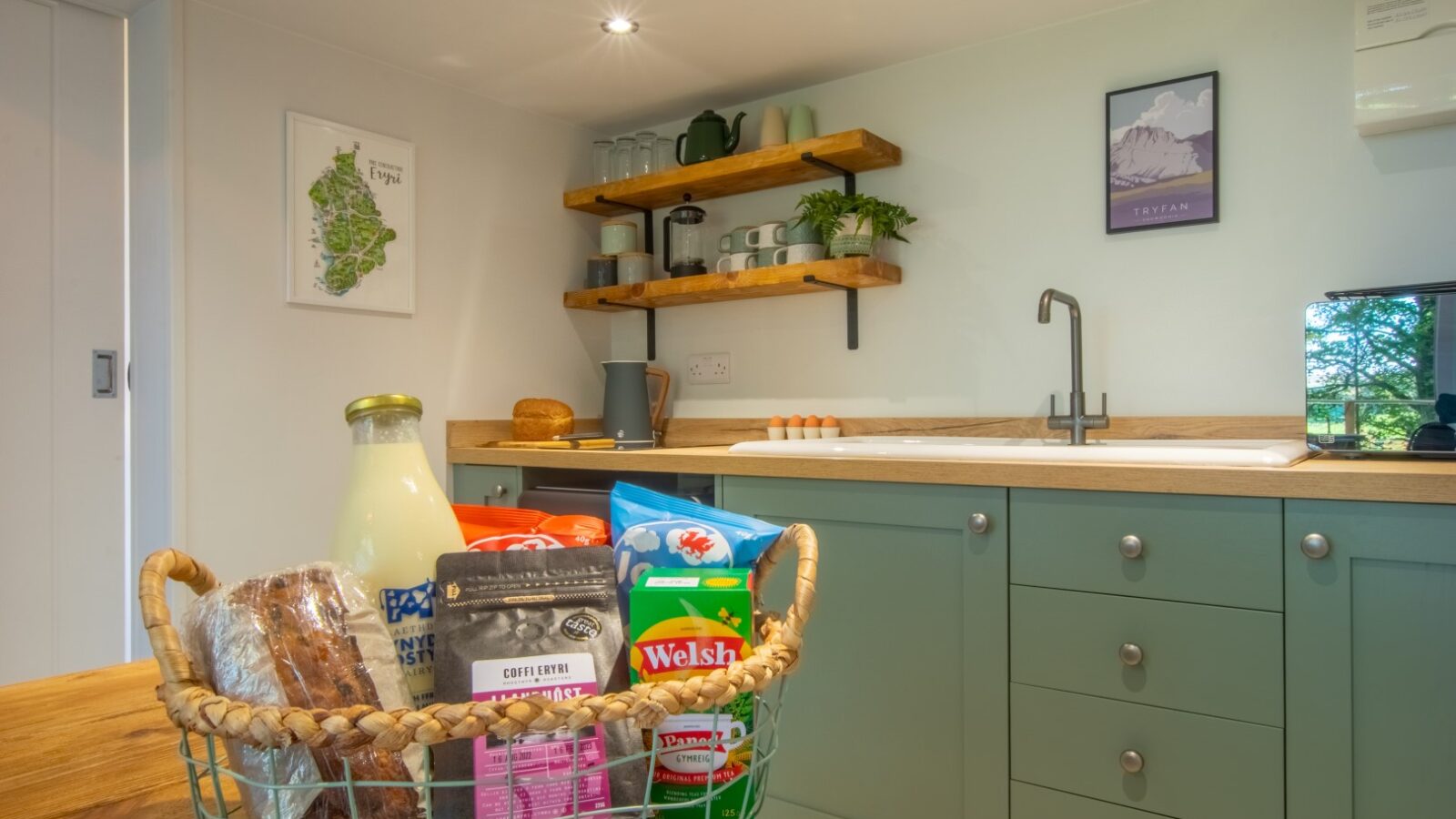 A kitchen reminiscent of a cozy glamping retreat, featuring green cabinets and wooden shelves. On the table, a basket of groceries—milk, bread, and snacks—adds a touch of Porth Eryri's rustic charm.