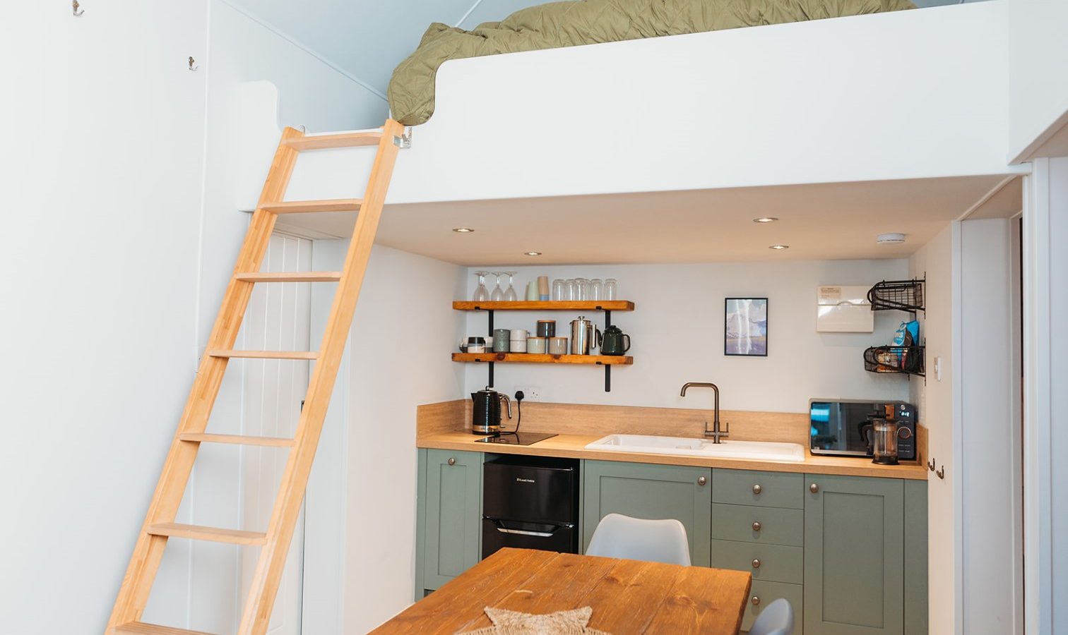 The compact kitchen and dining area, inspired by Porth Eryri's charm, features green cabinets, a wooden table, and a cozy loft bed accessible by a sturdy wooden ladder.