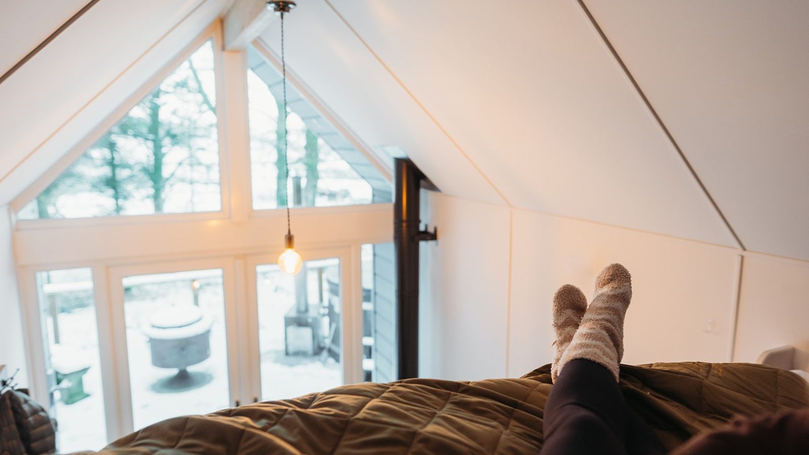 A person with socked feet relaxes in a cozy loft, gazing out at the snow-covered outdoors through large triangular windows reminiscent of Porth Eryri's breathtaking vistas.