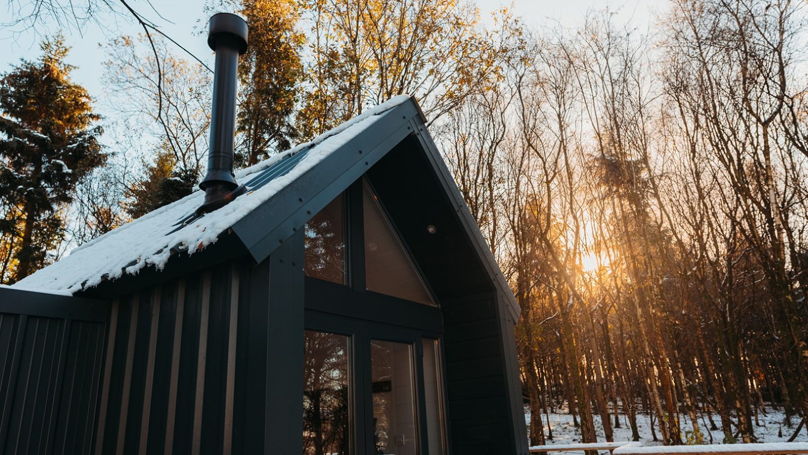 A small black cabin with a snow-covered roof stands in a forest near Porth Eryri, with the sun setting through the trees.