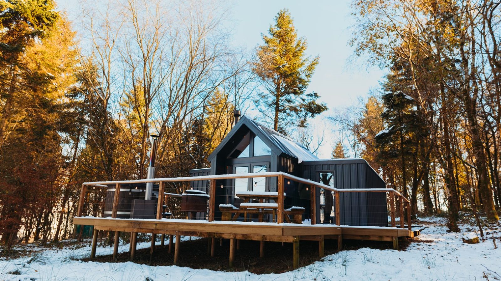 Nestled amidst the serene beauty of a snowy landscape, this A-frame cabin features a charming wooden deck. Surrounded by trees and basking under a clear blue sky, it's reminiscent of the tranquil charm found near Porth Eryri.