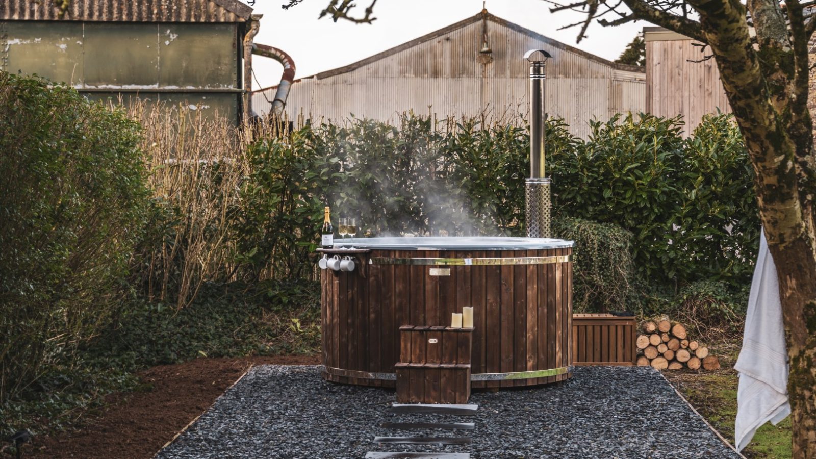 A rustic outdoor hot tub at Bogee Farm is set on a gravel platform with stepping stones leading to it. Steam rises from the water, surrounded by greenery and wooden sheds. A white towel hangs on a tree branch nearby, and firewood is stacked in the background.