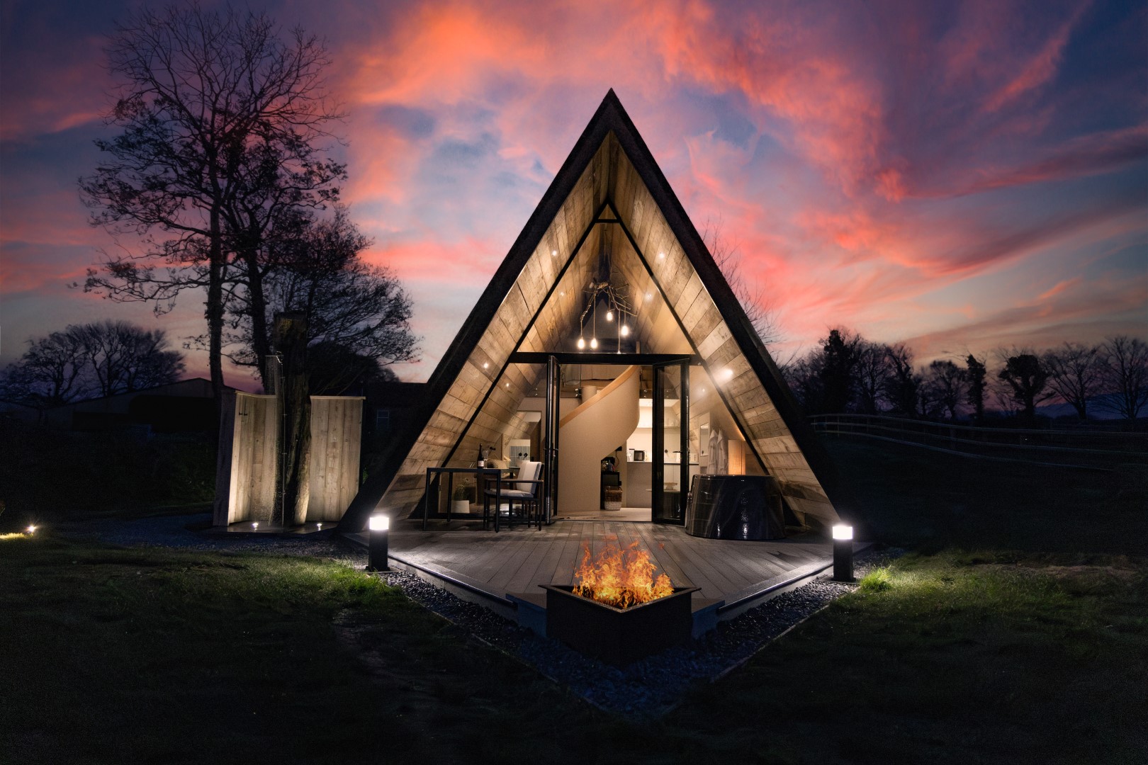 A-frame cabin with large windows, illuminated from within, facing a fire pit against a vibrant sunset sky—perfect for a romantic Christmas break for two in the UK. The Tri-pod at Pantcefn Glamping