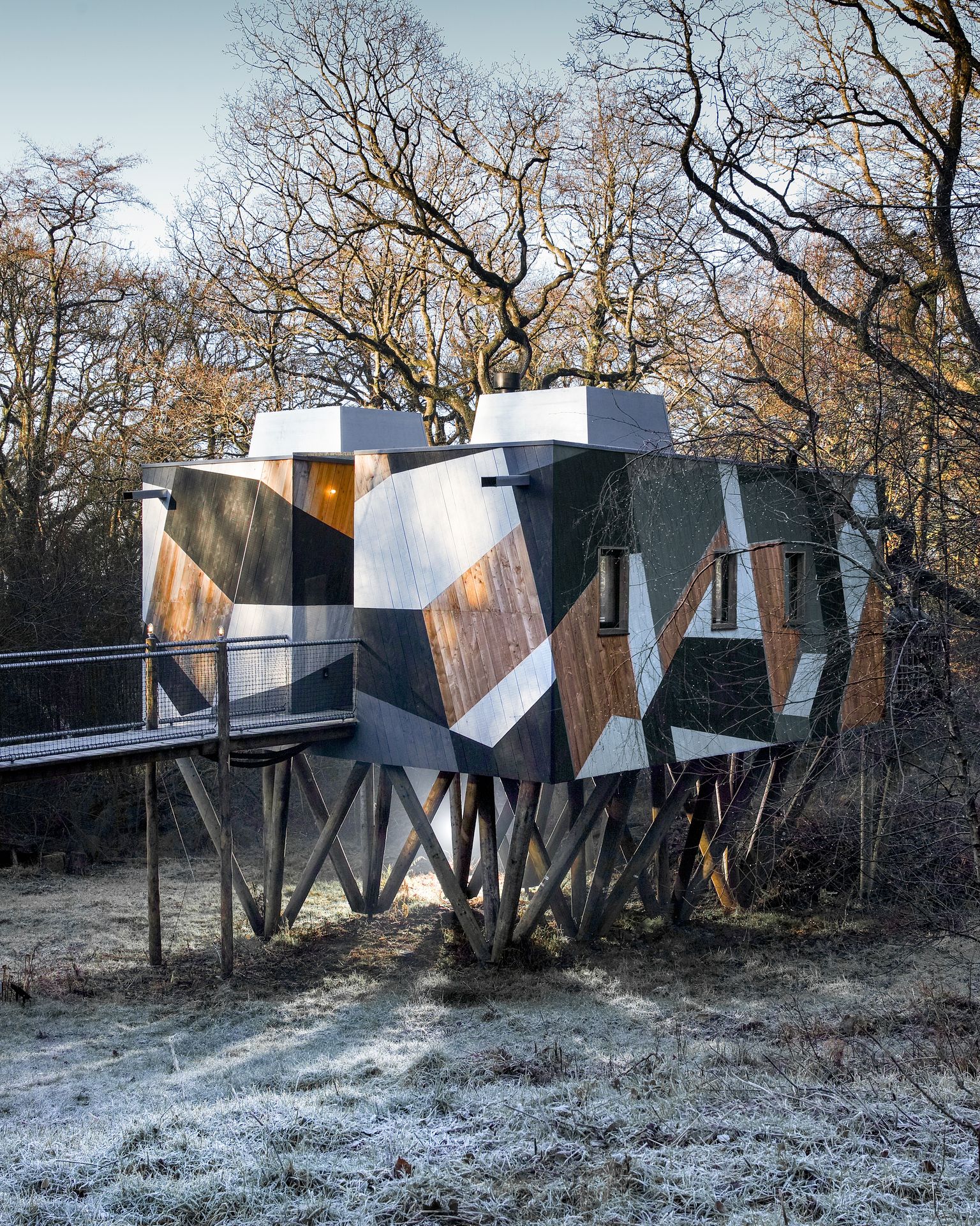 A modern, geometric treehouse with a camouflage design stands on stilts in a frosty, leafless forest setting, making it one of the best Xmas breaks in the UK.