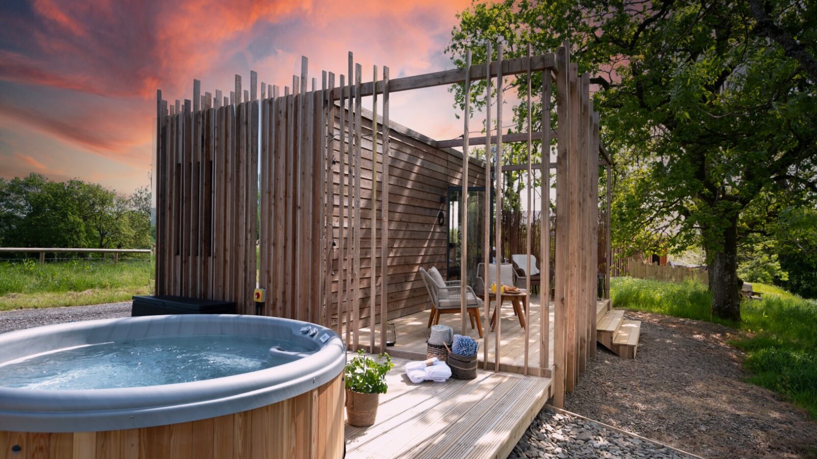 Modern wooden cabin with an outdoor hot tub on a deck, set against a vibrant sunset sky and surrounded by greenery. Experience tranquility at Pond View Lodges, where nature's beauty captivates.