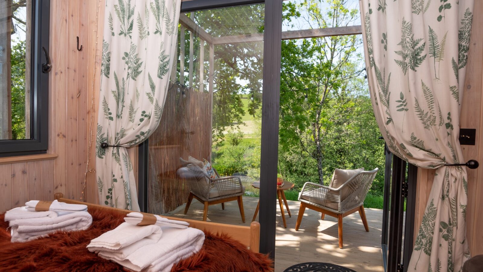 Sunny room with fern-patterned curtains, a glass door leading to a deck with two chairs, and folded towels on a fur-covered bed. Enjoy the tranquil view of a nearby pond, perfect for moments of relaxation. 