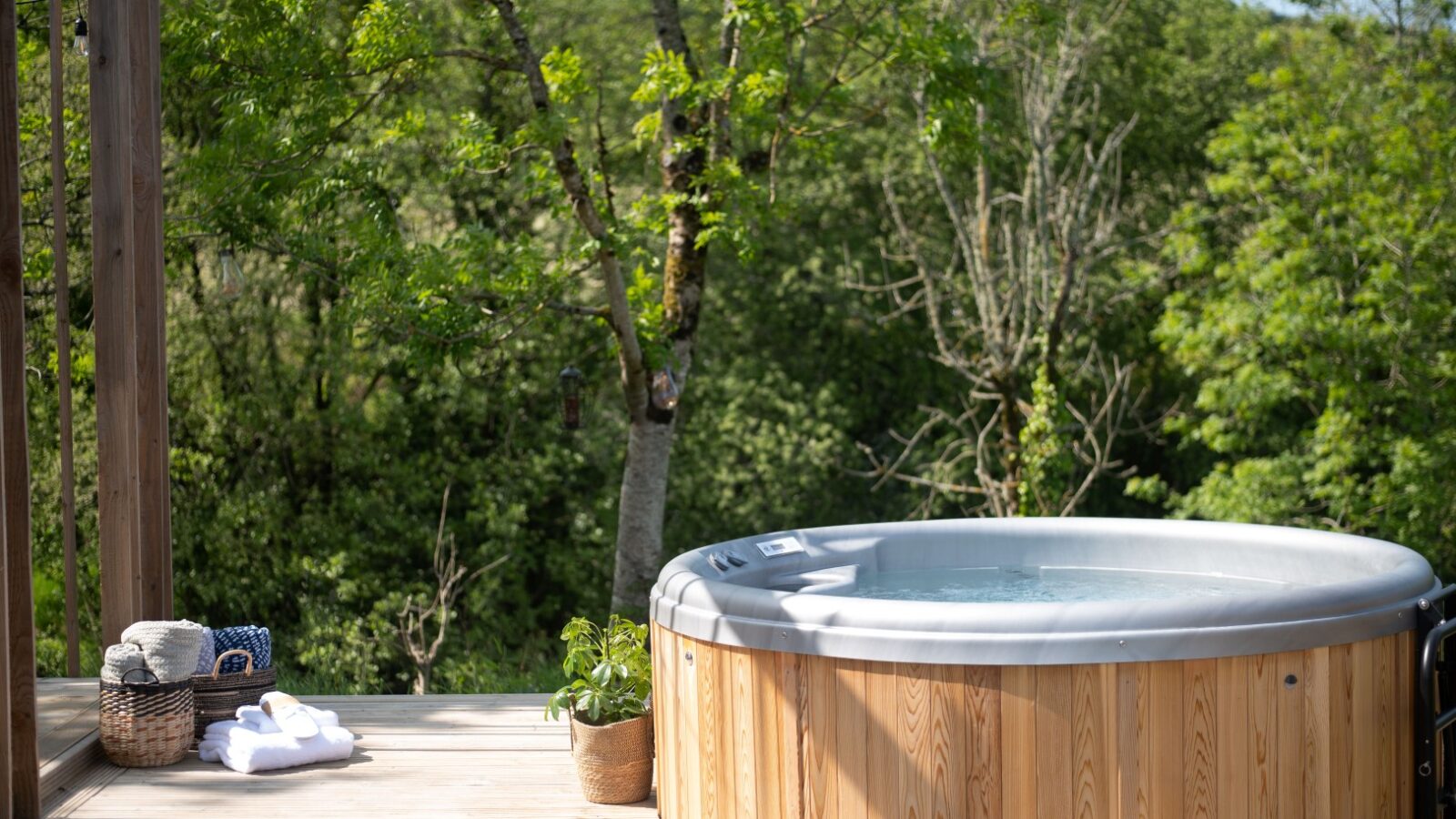 A wooden hot tub on a deck overlooks the serene Pond View, surrounded by lush green trees. Nearby, a basket of towels and a small potted plant enhance the tranquil ambiance, making this setting perfect for those seeking relaxation at Pond View Lodges.