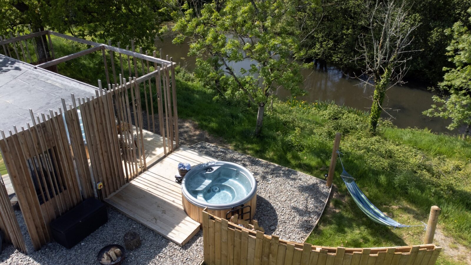 Aerial view of a wooden deck with a round hot tub, nestled among trees and overlooking the tranquil waters of Pond View Lodges. Nearby, a hammock sways gently on the grass by the river.