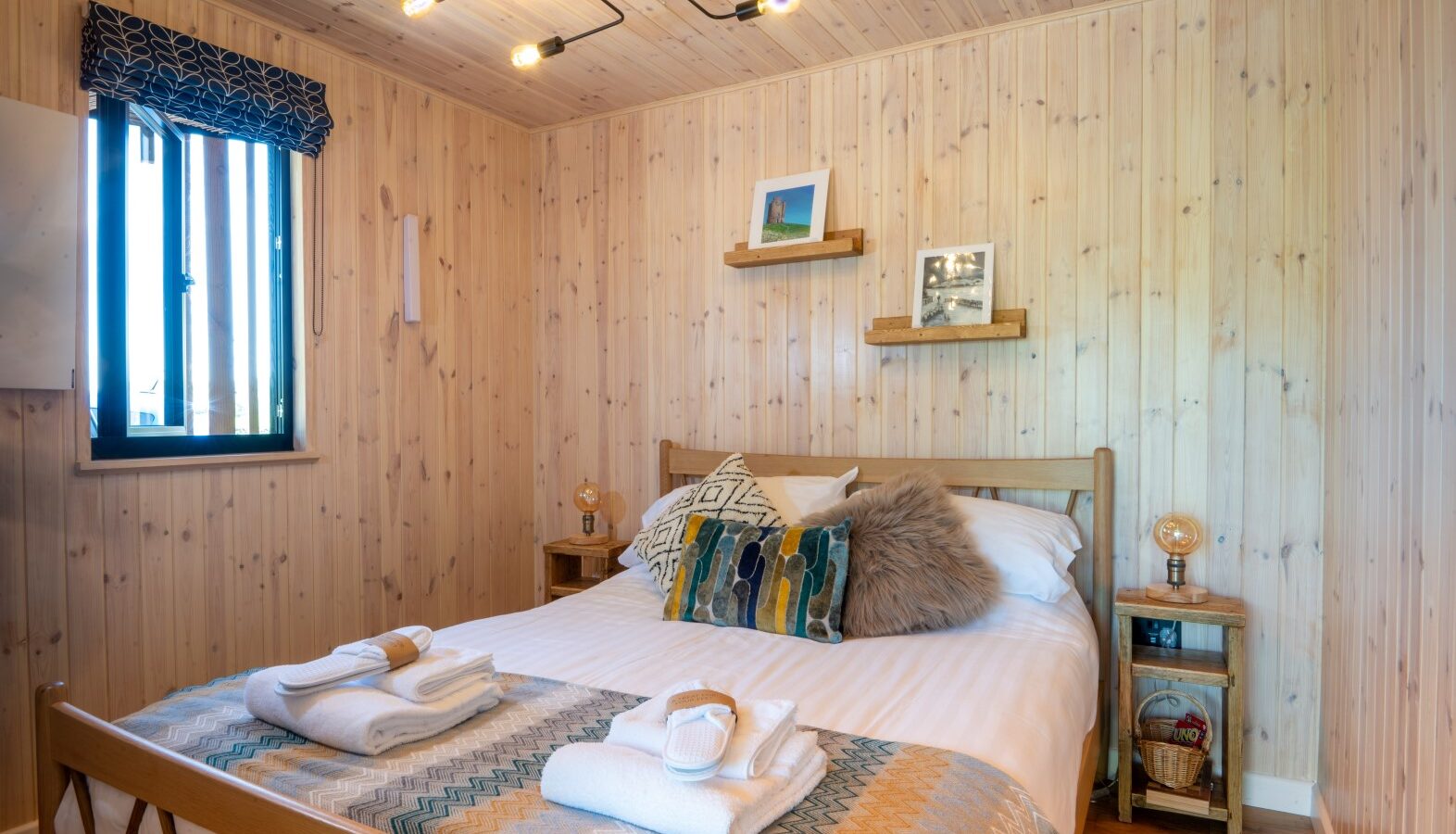 Cozy bedroom with wood-paneled walls, a neatly made bed, colorful pillows, lamps on nightstands, and framed photos on shelves, capturing the charm of Pond View Lodges.