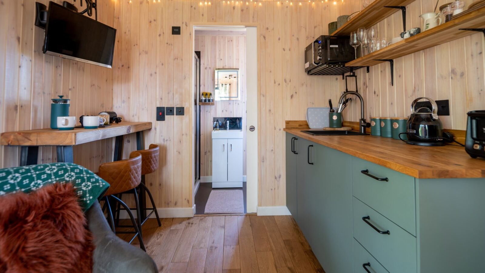 Compact modern kitchen at Pond View Lodges features green cabinetry, wooden countertops, a wall-mounted TV, and string lights. A doorway leads to a bathroom.