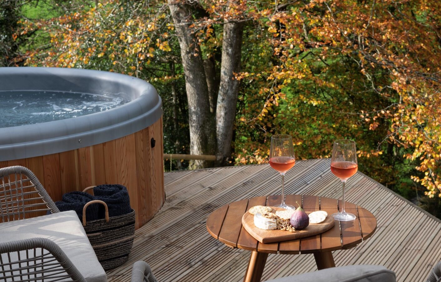 Outdoor deck overlooking picturesque Pond View Lodges, complete with a hot tub, surrounded by autumn trees. A table holds two glasses of rosé wine, alongside cheese and a fig, offering a perfect retreat.