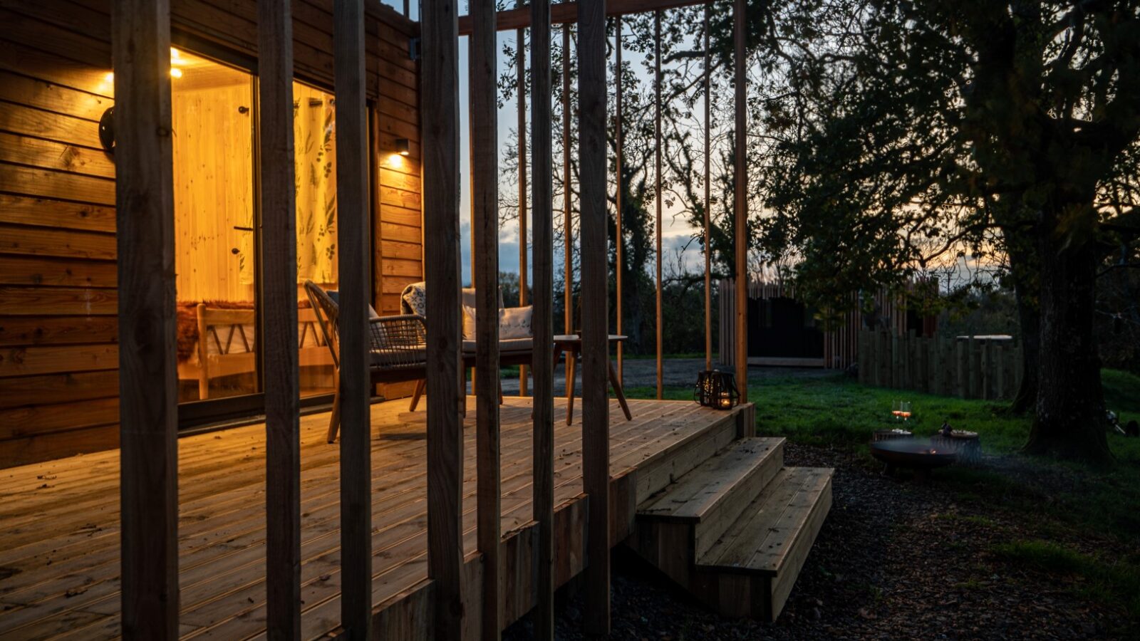 Wooden cabin with an illuminated porch at dusk, surrounded by trees and featuring wooden steps and outdoor seating, offering a serene Pond View.