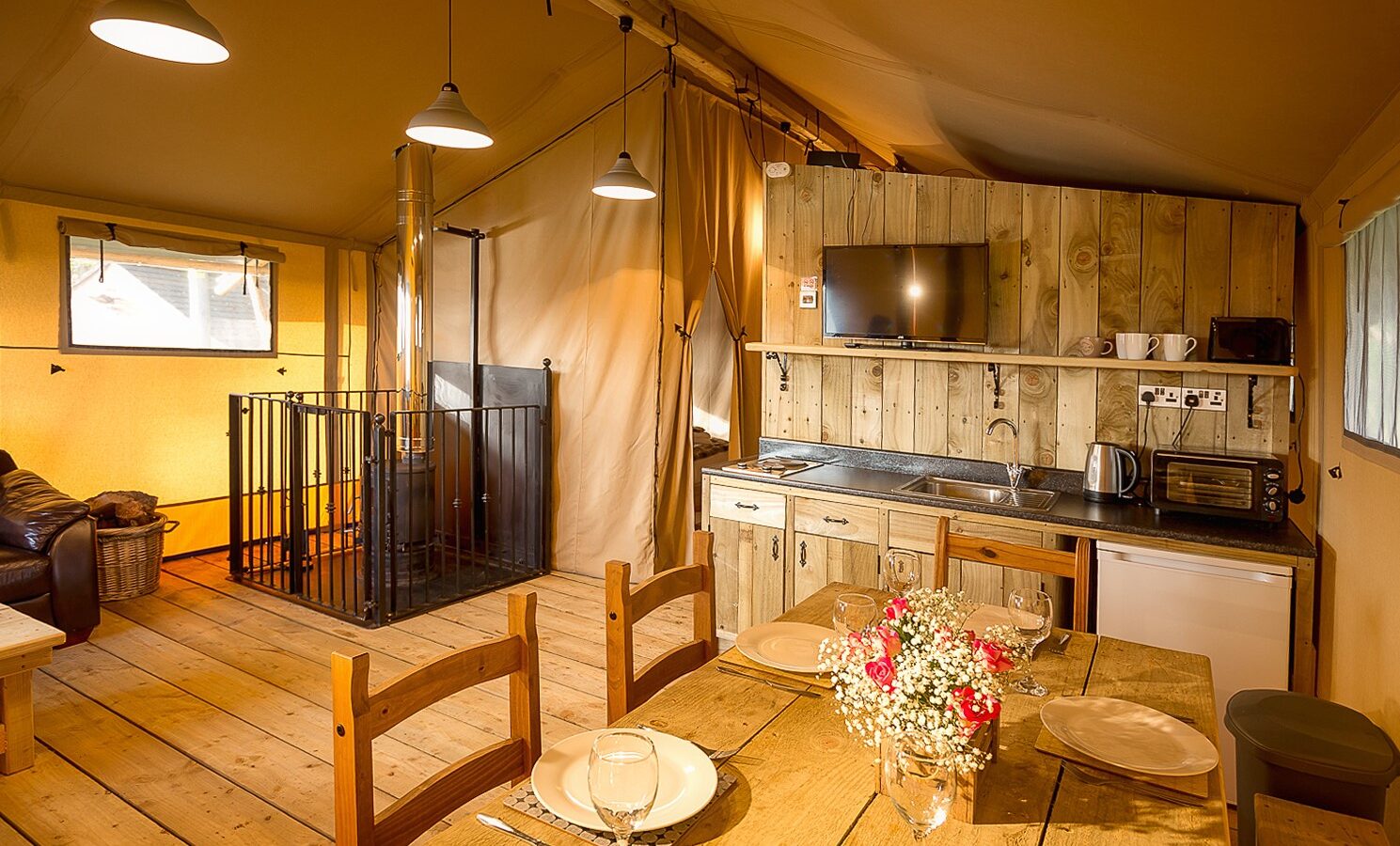 The interior of the rustic cabin at Sweeney Farm features wooden furniture, a cozy dining table set, and a quaint kitchenette. A wood-burning stove in the corner adds to the glamping charm, providing warmth and ambiance for an unforgettable stay.