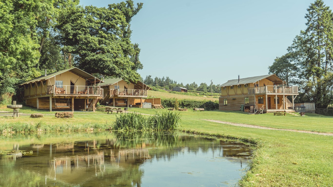 Experience the charm of Sweeney Farm, where glamping tents and cabins nestle beside a tranquil pond, all set in a grassy landscape adorned with trees under a clear sky.