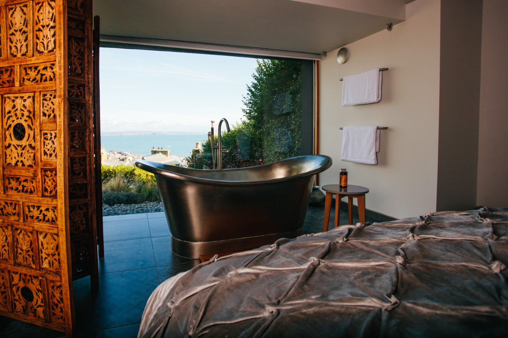 A luxurious bathroom in one of the best dog-friendly places to stay in Cornwall, featuring a freestanding tub, ornate wooden screen, and a scenic ocean view through an open wall of windows. Perfect for those seeking holiday cottages near Eden Project.