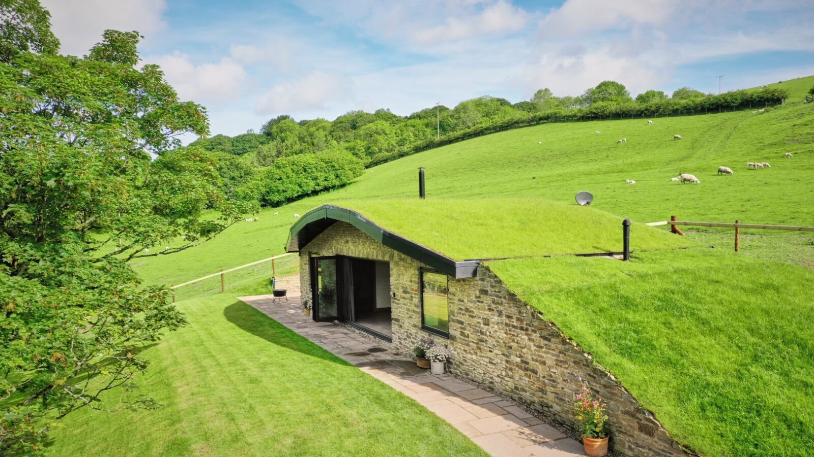 The Dolassey hillside house, resembling a cozy burrow, features a grass-covered roof and stone walls. It's nestled along a path flanked by green fields and trees beneath the expansive blue sky.