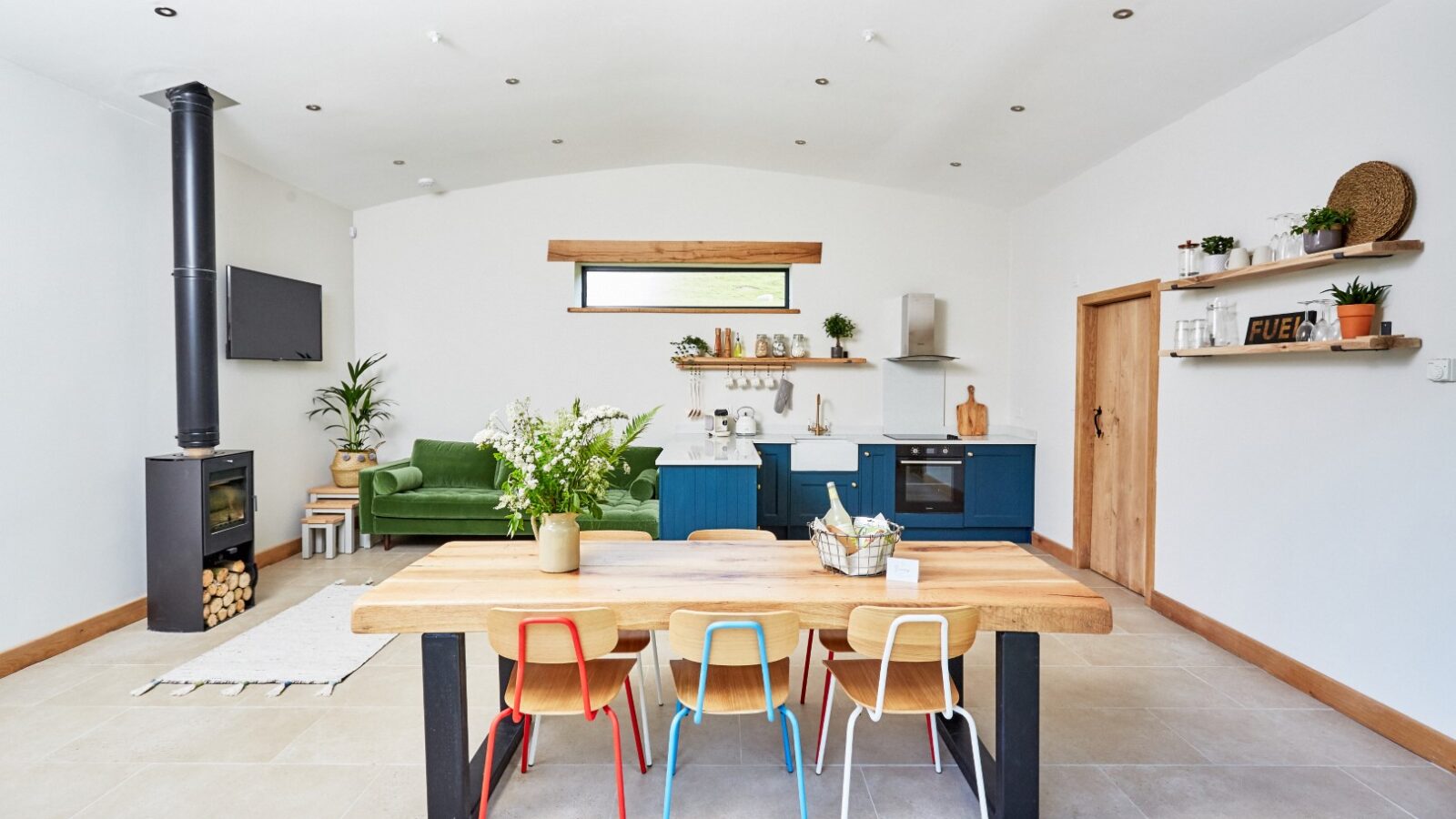 Modern open-plan kitchen and living area with a Burrows Dolassey dining table, colorful chairs, green sofa, and wall-mounted TV.