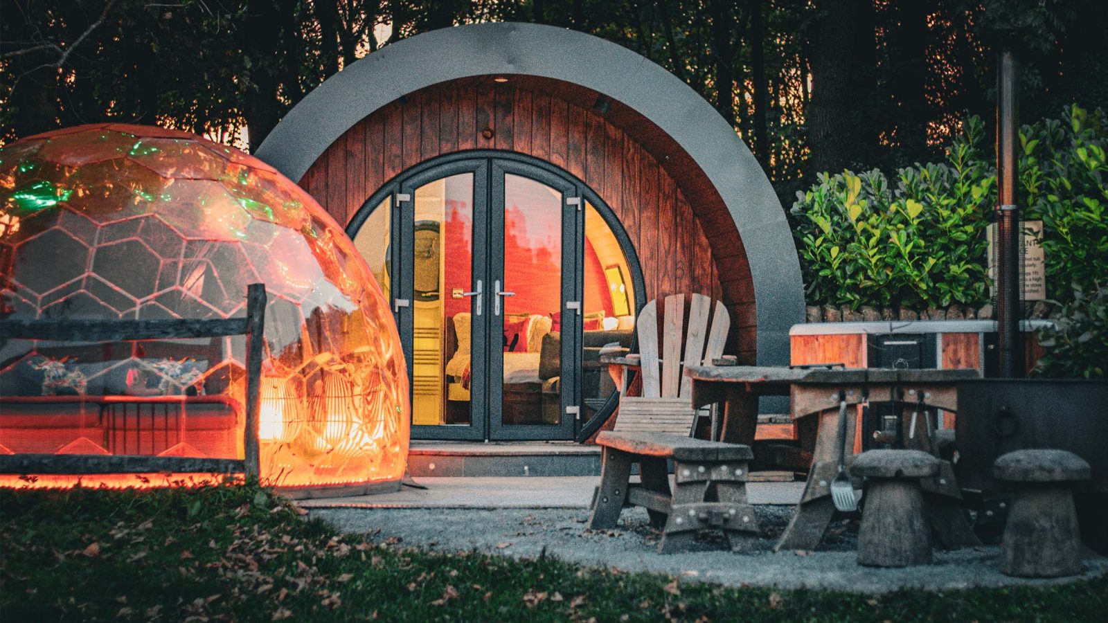 Dome-shaped cabin at Catgill Farm with a transparent bubble dome and wooden chairs in a wooded area at dusk.