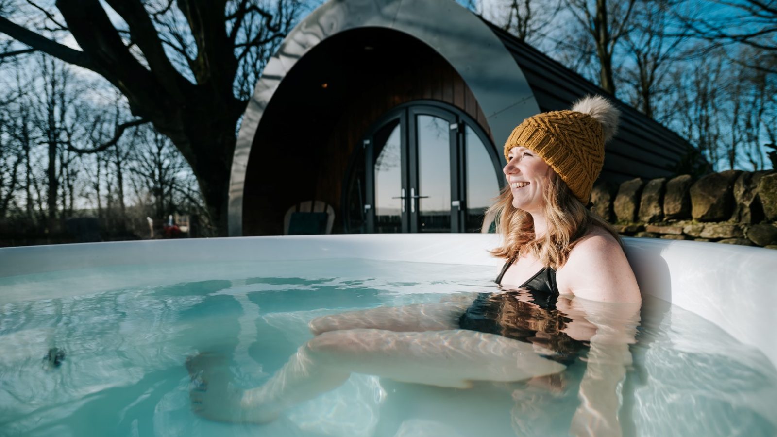 A person in a yellow beanie relaxes in an outdoor hot tub at Catgill Farm, with a curved cabin and leafless trees behind.