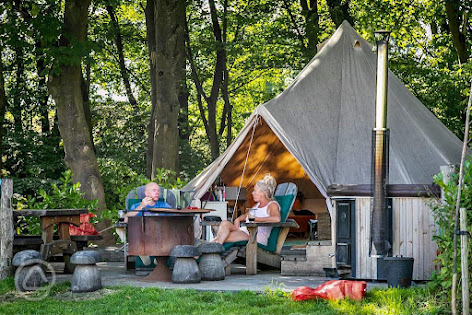 Two people sit outside a canvas tent at Catgill Farm, surrounded by trees, enjoying the outdoors with wooden tables and chairs.