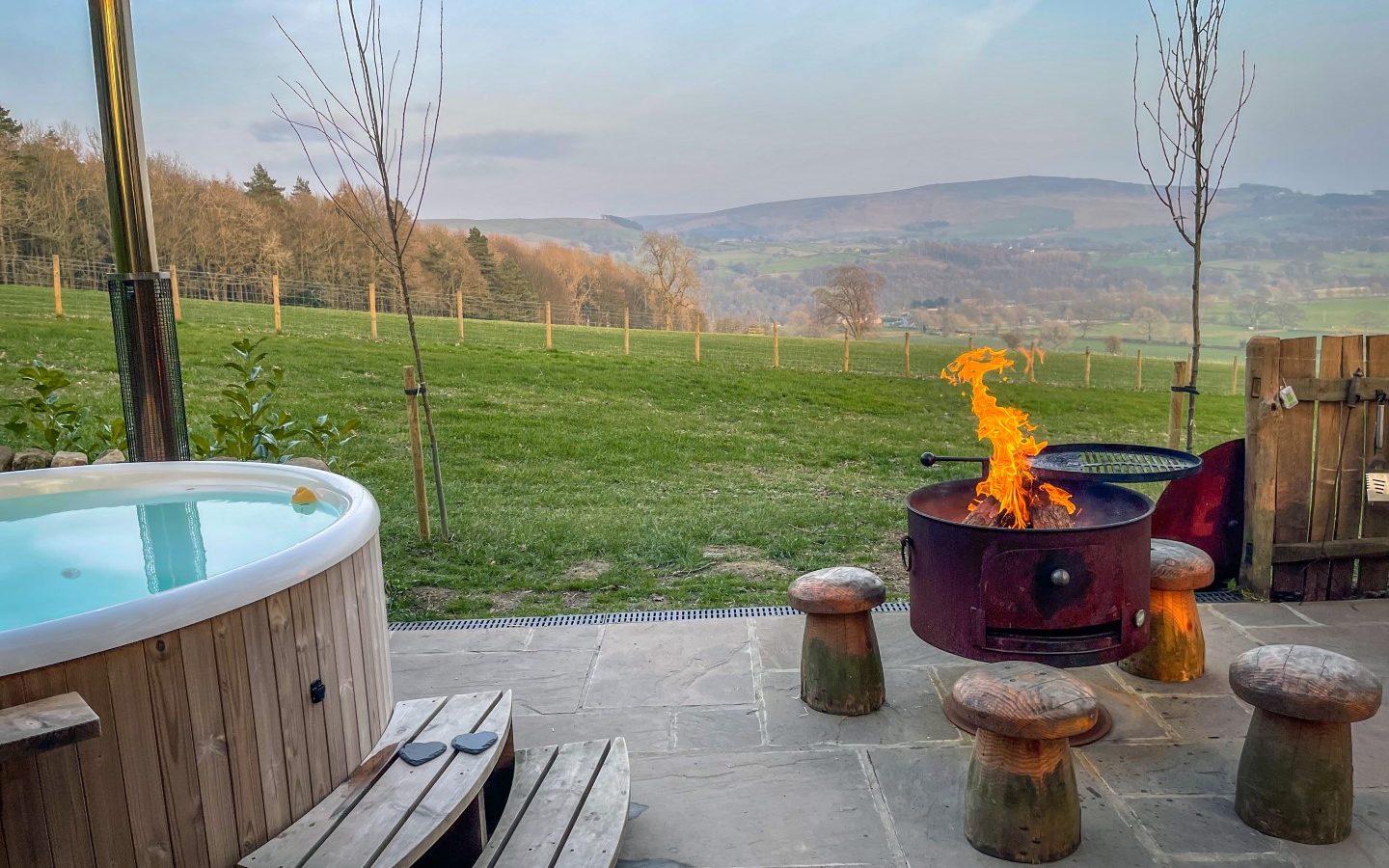 Catgill Farm's wooden hot tub and fire pit with stools offer a serene view of the grassy field and rolling hills.