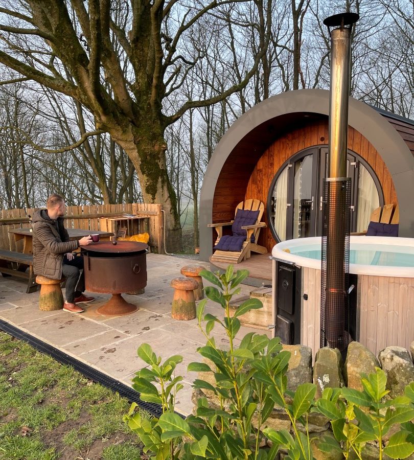 A person relaxes at a patio table next to a fire pit in front of a modern wooden pod cabin at Catgill Farm. A hot tub and outdoor chairs provide comfort amid the wooded area, where trees beautifully frame the grassy lawn.