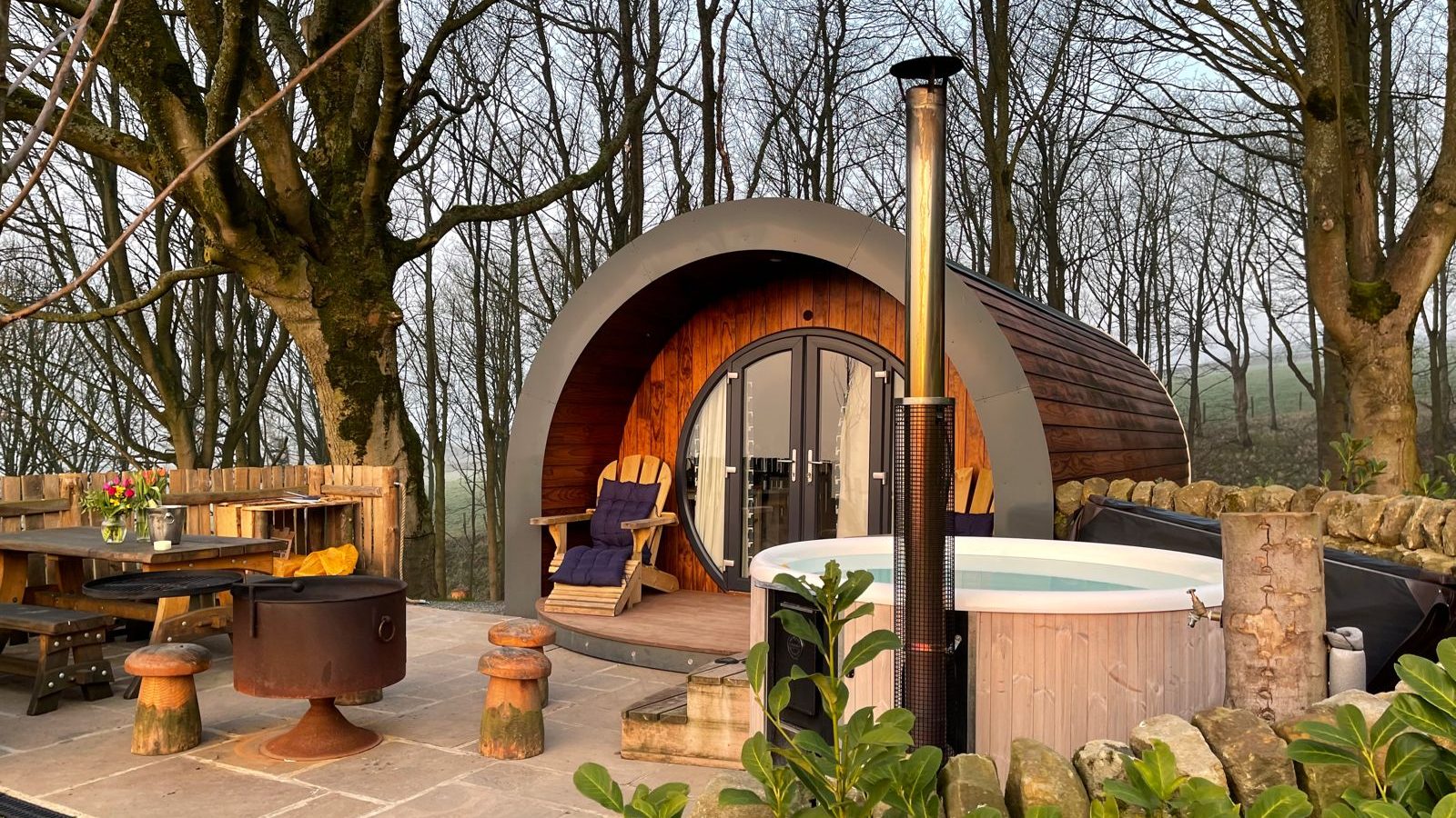 A cozy wooden cabin with a curved roof nestled in a forested area of Catgill Farm. The patio features a hot tub, outdoor seating with cushions, a picnic table, and a fire pit. Bare trees surround the area, and early morning light creates a serene atmosphere perfect for a Yorkshire Dales farm stay.