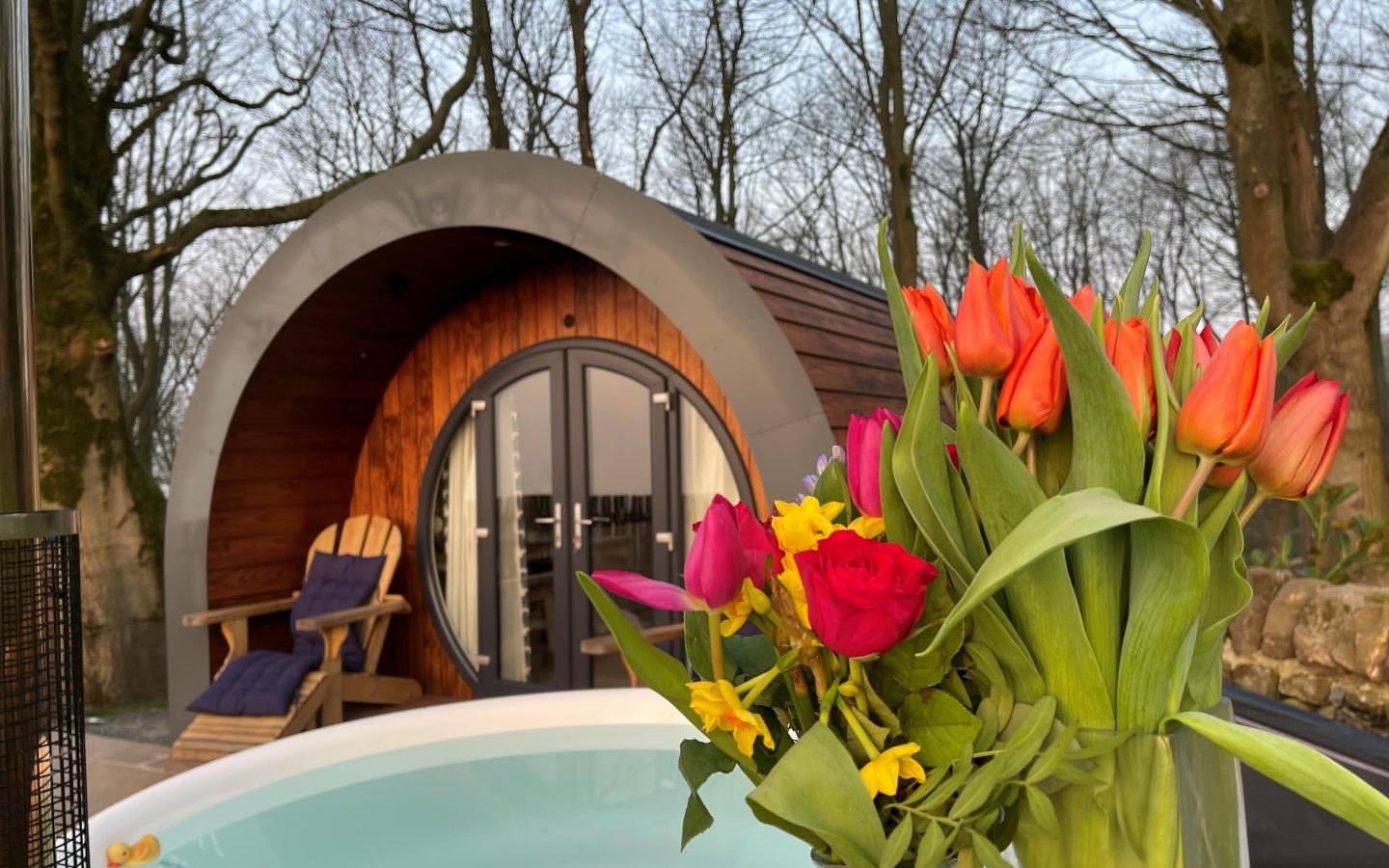 A cozy wooden cabin with a curved roof nestled in the forested area of Catgill Farm. In the foreground, there's a hot tub and a vibrant bouquet of orange tulips, red roses, and yellow flowers. Two wooden chairs are placed invitingly in front of the cabin.