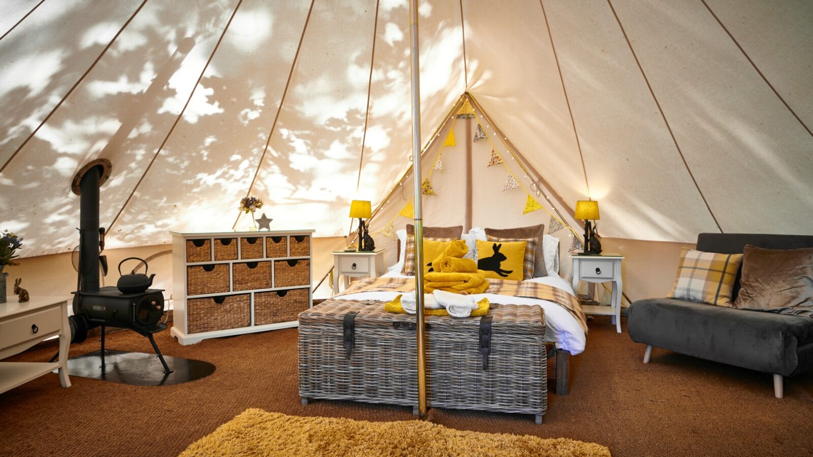 Inside a cozy glamping tent at Catgill Farm in the Yorkshire Dales, you'll find a bed, wicker trunk, dresser, sofa, and a small wood stove. Decor includes charming yellow accents for a delightful farm stay experience.