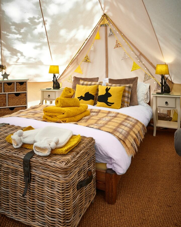 Experience the cozy charm of a glamping tent at Catgill Farm, featuring a comfortable bed, yellow and brown accents, a wicker chest, bedside tables, and decorative pillows.