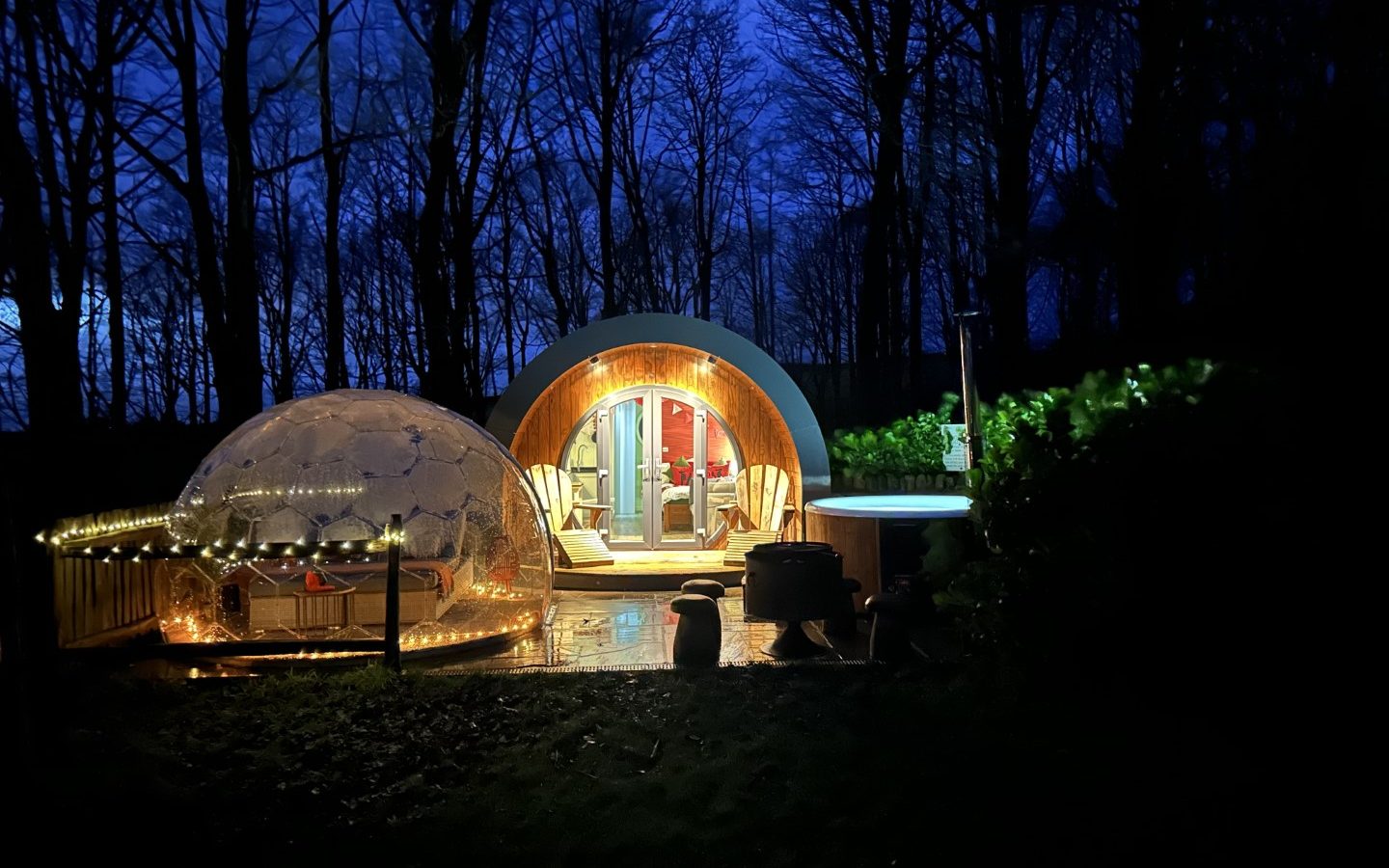 A cozy dome-shaped cabin with warm lights sits amidst the serene Yorkshire Dales at dusk. At Catgill Farm, a transparent igloo adorned with string lights complements the scene, featuring a hot tub on a wooden deck nestled among the trees for a perfect farm stay experience.