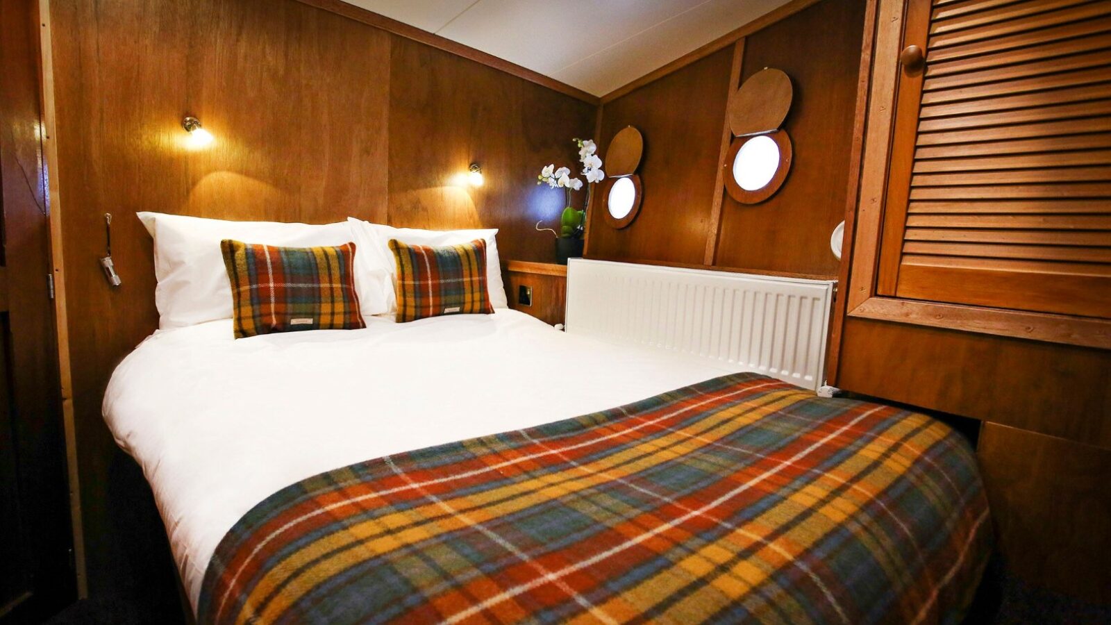 Cozy cabin bedroom with wooden walls, plaid bedding reminiscent of Argyll Cruising, two portholes, and warm lighting.