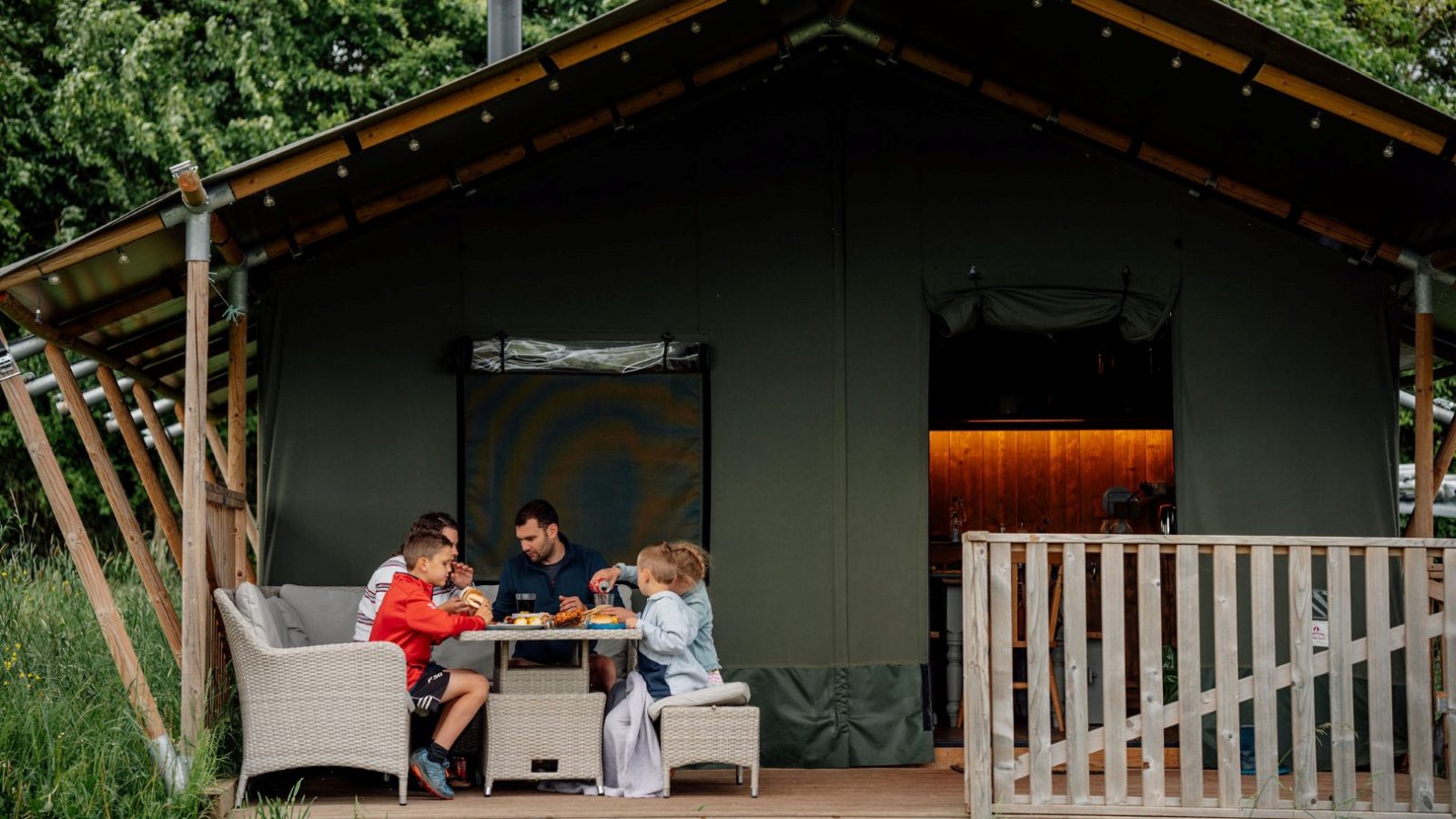 A family is seated around a table outside their Nature's Nest tent, enjoying a meal. Set on a wooden deck in a grassy area, with trees filling the background, this glamping scene conveys a relaxed, outdoor ambiance.