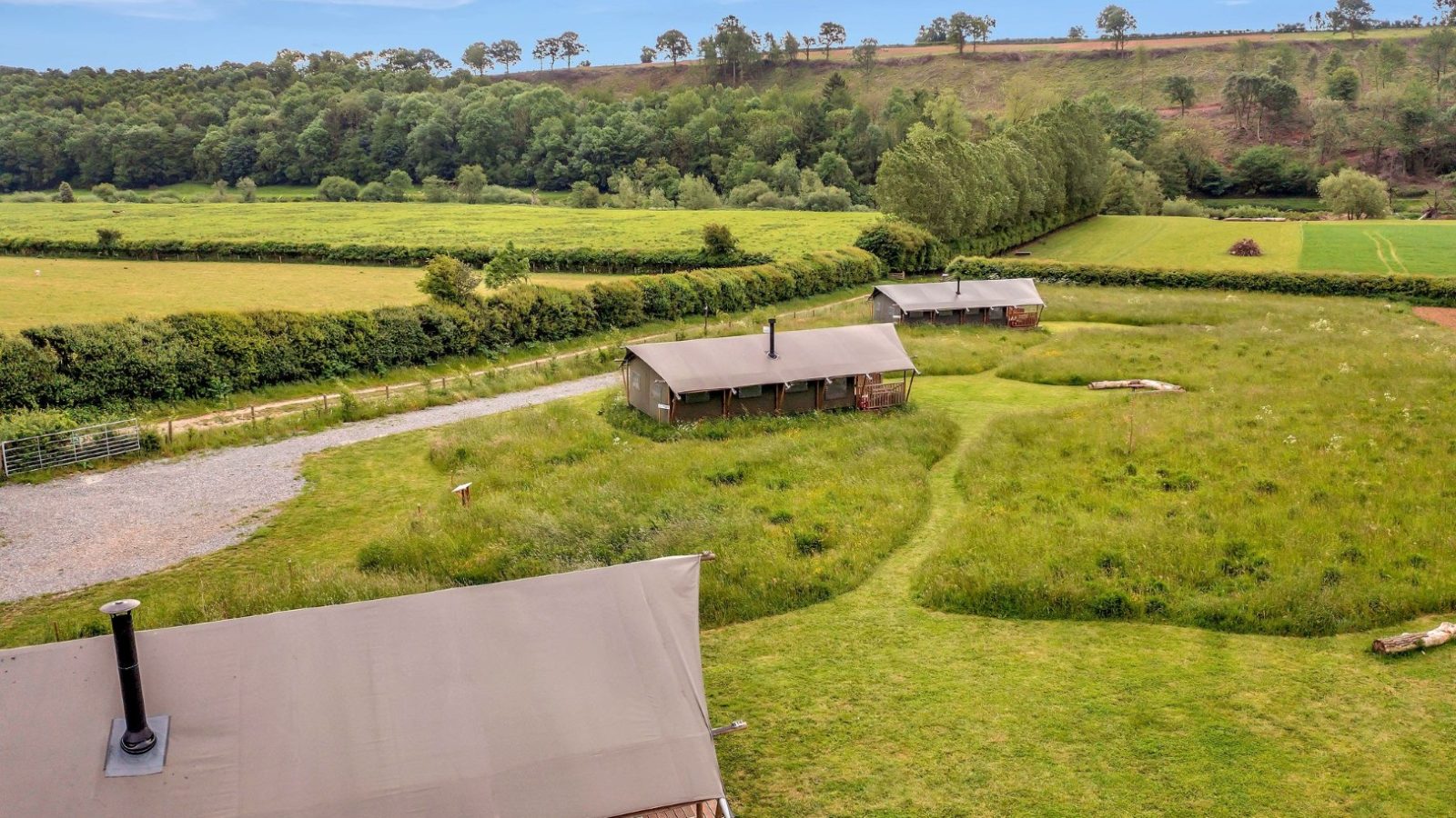 An aerial view of Nature's Nest Glamping reveals two large tents nestled in the lush, green countryside. Surrounded by grassy fields and trees, a gravel path winds through this serene landscape, set against a wooded hill under a clear blue sky.