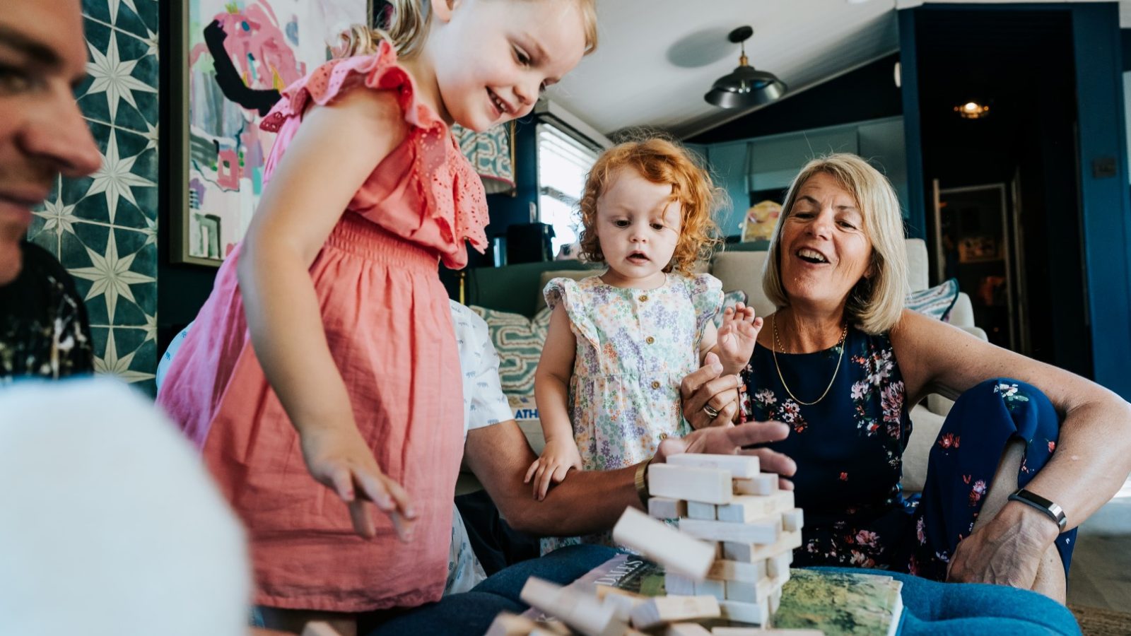 A young girl knocks over a tower of wooden blocks at Howgill Lodge while a toddler and two adults look on, smiling.