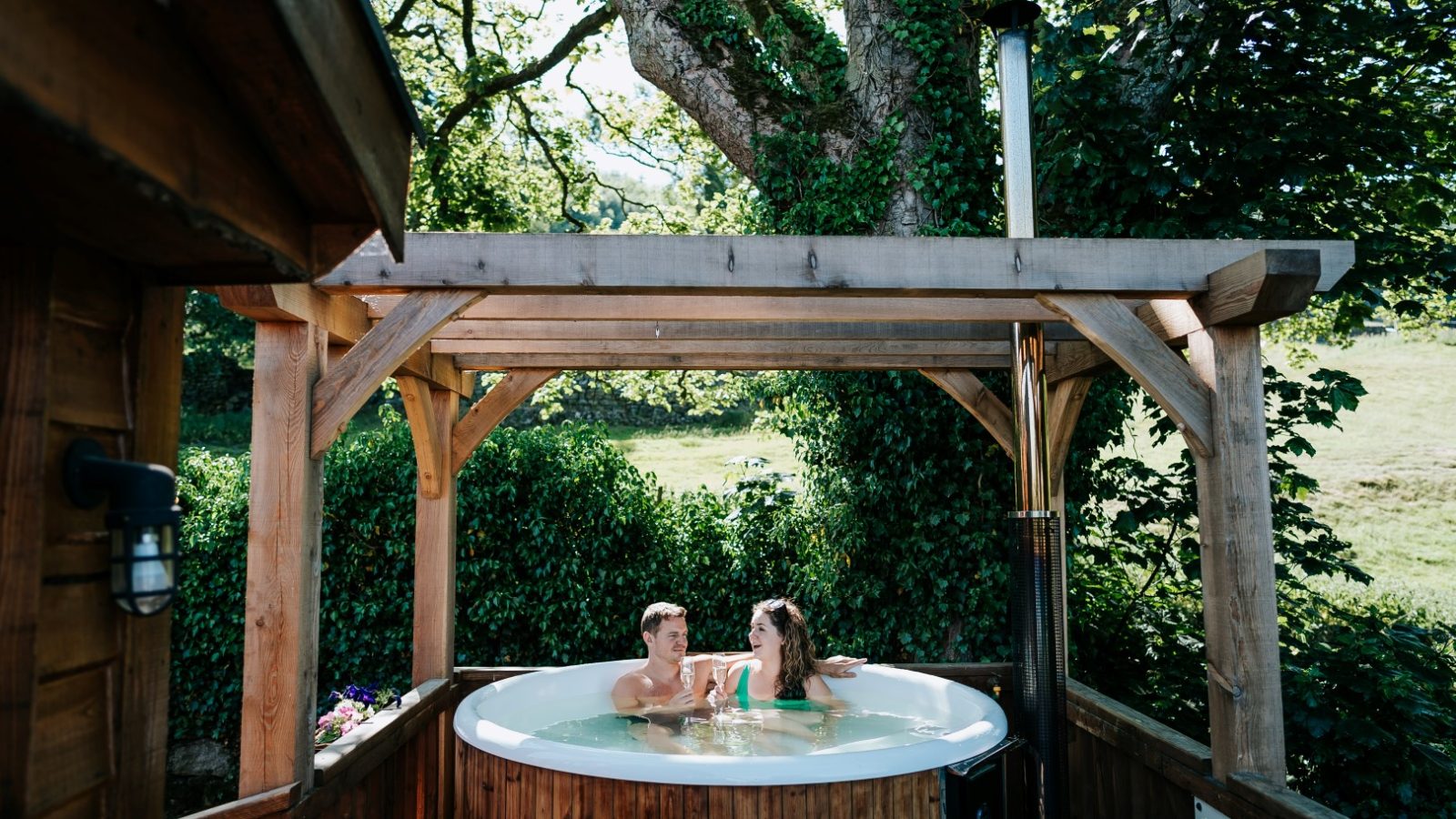 Two people relax in a wooden hot tub under a pergola at Howgill Lodge, enveloped by lush greenery and trees.