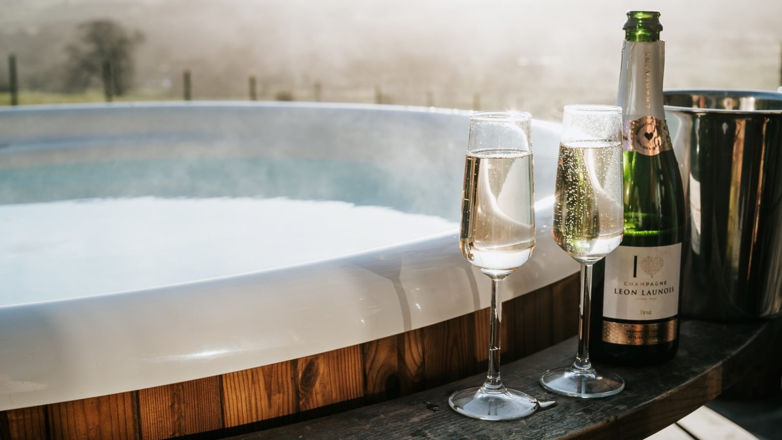 Two glasses of champagne and a bottle beside a hot tub at Howgill Lodge, with a scenic view in the background.