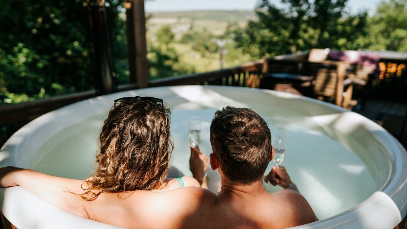 Two people relax in an outdoor hot tub at Howgill Lodge, champagne in hand, surrounded by lush greenery and distant hills.