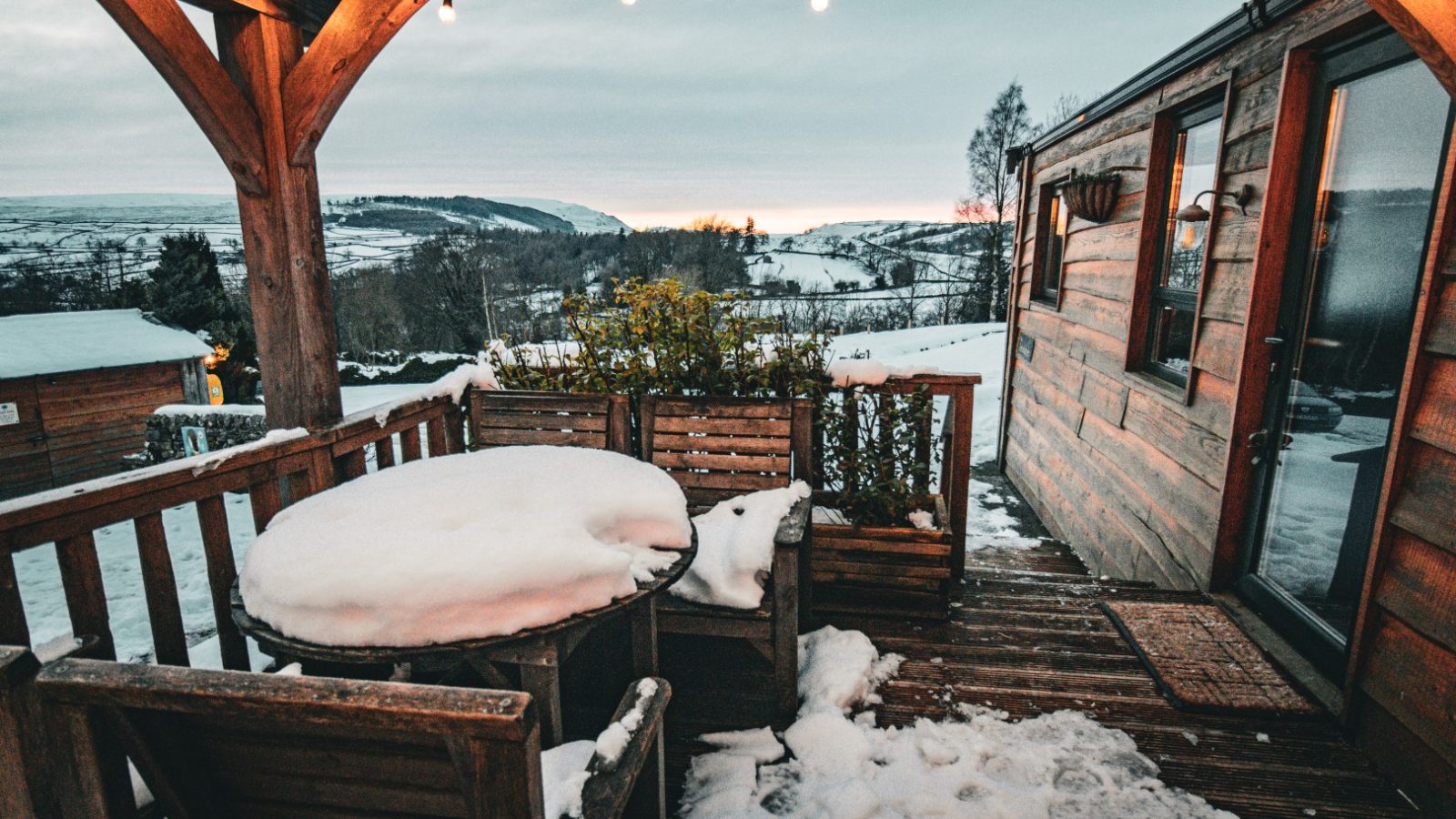 Snow-covered patio at Howgill Lodge has wooden furniture under string lights, overlooking a winter landscape with mountains.