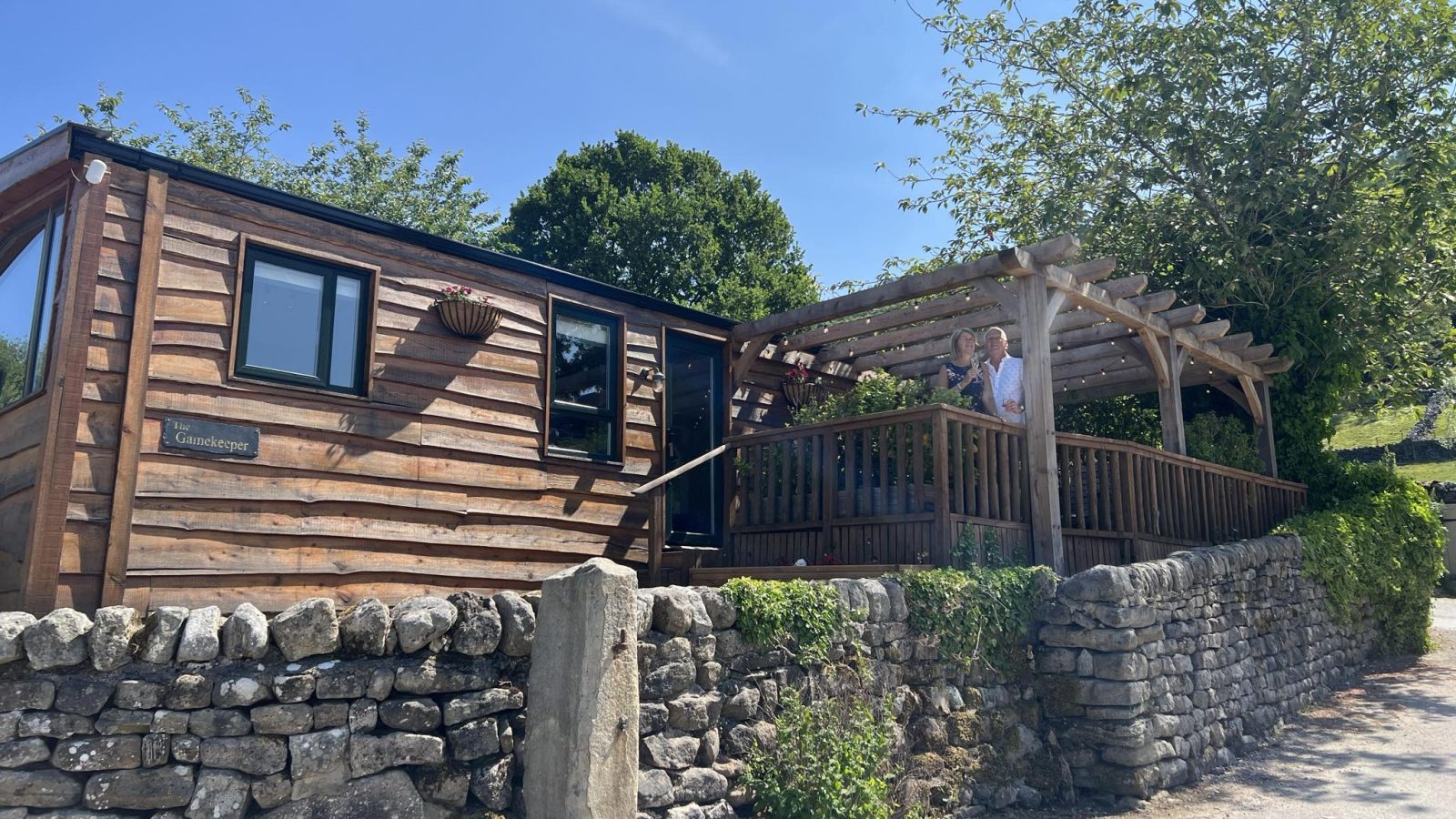 Wooden cabin at Howgill Lodge with a stone wall and pergola. Two people stand on the deck, surrounded by trees under a blue sky.