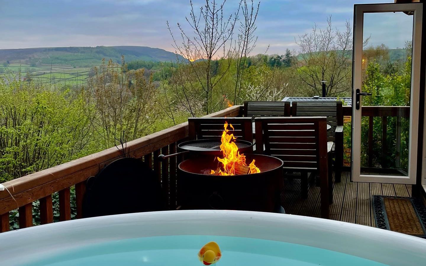 A deck at Howgill Lodge features a hot tub, lit fire pit, table, chairs, and open door overlooking a scenic hilly landscape.