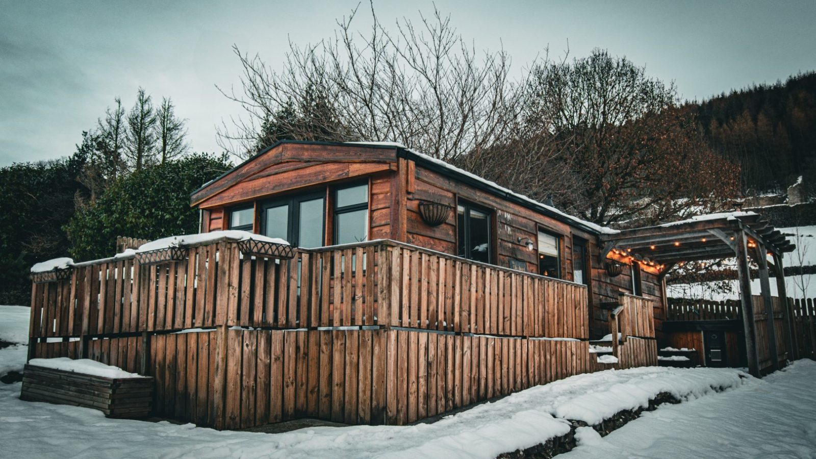 Howgill Lodge: A wooden cabin with a fenced deck on a snowy landscape, surrounded by trees under an overcast sky.