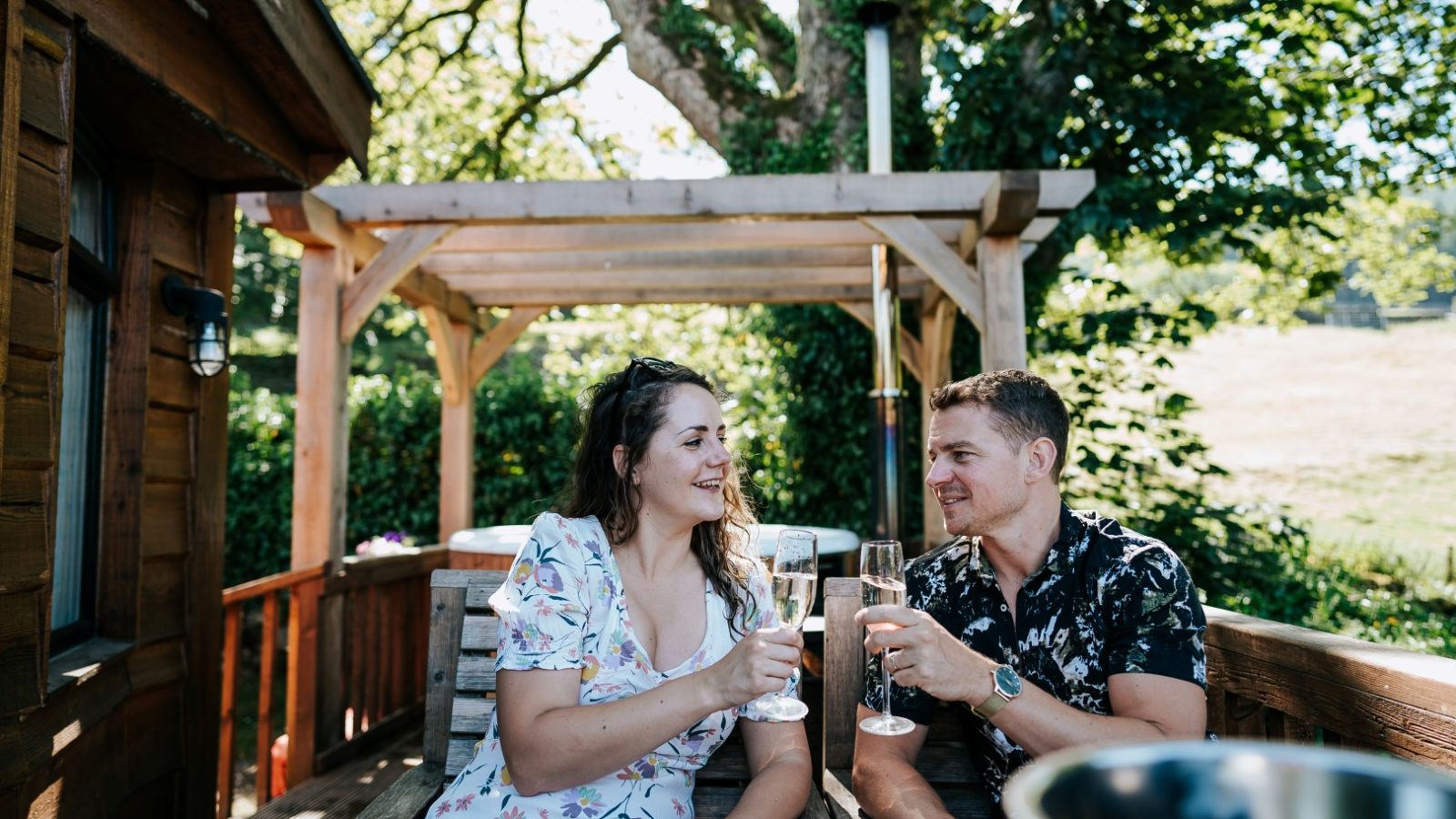 A man and woman sit outdoors on a wooden bench under a pergola at Howgill Lodge, clinking champagne glasses and smiling.